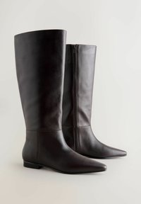 Ett par höga, mörkbruna läderstövlar med spetsiga tår och siddragkedjor, visade mot en enkel ljus bakgrund.
