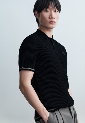 Jeune personne aux cheveux noirs courts portant un polo noir à manches courtes côtelées et un pantalon gris plissé, debout avec les mains dans les poches.