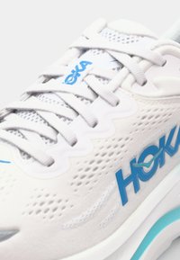 Vit idrottssko med mesh-detaljer, blå logotyp och grå snören. Har en tjock sula med en ljus mintfärgad accent för dämpning.