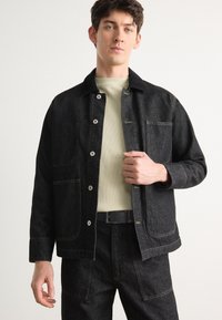 Giacca di jeans nera con due tasche frontali, bottoni argentati e cuciture in vista. Indossata sopra una camicia chiara e strutturata e pantaloni neri abbinati.