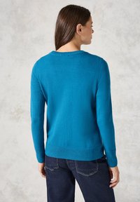 Pull en tricot bleu sarcelle à manches longues, avec un col et un ourlet côtelés, associé à un jean foncé. Texture douce et design ajusté.