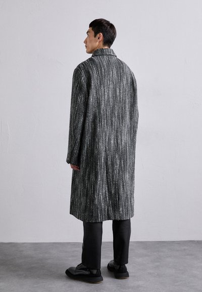 Manteau gris et noir à motifs avec une coupe longue et droite, un large col et un tissu texturé, associé à un pantalon et des chaussures noires.