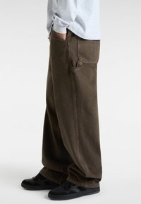 Stora, mörkbruna byxor med en smidig yta och sidofickor, kombinerade med svarta sneakers, står på en ljus bakgrund.