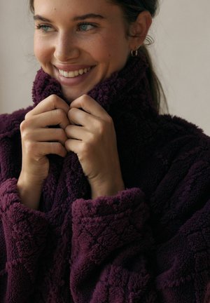 Plush lila fleece-plagg med hög krage, med en texturerad yta. Modellen håller i kragen och visar upp en mysig design.