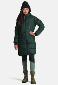 Lang groene pufferjas met een rits aan de voorkant, kinbeschermer en zijzakken. Gepaard met groene legging en olijfkleurige laarzen. Voorzien van een gebreide beanie.