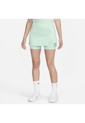 Frau trägt ein mintgrünes sportliches Skort mit Shorts darunter, weißen Nike-Socken und weißen Nike-Sneakern, steht vor weißem Hintergrund.