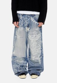 Φαρδιά ξεθωριασμένα μπλε τζιν με λευκά σχέδια αγγέλων και αλυσίδας, φορεμένα με μαύρα αθλητικά παπούτσια και μαύρο μακρυμάνικο τοπ.