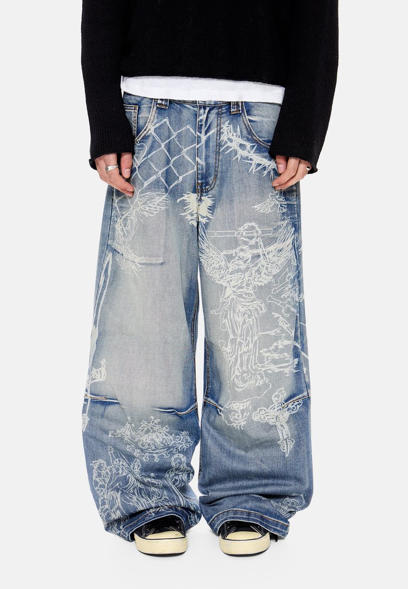 Φαρδιά ξεθωριασμένα μπλε τζιν με λευκά σχέδια αγγέλων και αλυσίδας, φορεμένα με μαύρα αθλητικά παπούτσια και μαύρο μακρυμάνικο τοπ.