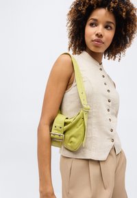 Γυναίκα με σγουρά μαλλιά φοράει αμάνικο μπεζ τοπ με κουμπιά, μπεζ παντελόνι και κρατάει μικρή lime πράσινη υφασμάτινη τσάντα ώμου με λουράκι και πόρπη.