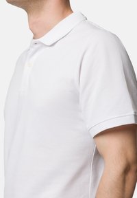 Biała koszulka polo z klasycznym kołnierzykiem, krótkimi rękawami i teksturowanym splotem; ma jeden guzik przy szyi.