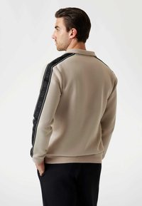 Felpa beige con strisce laterali nere caratterizzata da un motivo logo ripetuto. Tessuto morbido con colletto e polsini a costine.