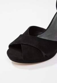 Sandale femme en daim noir à bout ouvert avec brides croisées et petit talon compensé sur fond blanc.