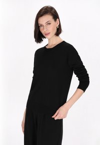 Zwarte langslieve trui met een ronde halslijn en ribbels aan de manchetten en onderkant. Zachte, gladde textuur; heeft een relaxte fit.