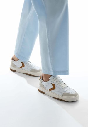 Sneaker bianchi e beige con suola in gomma testurizzata, dettagli beige chiaro e design con lacci, abbinate a pantaloni azzurri chiari.