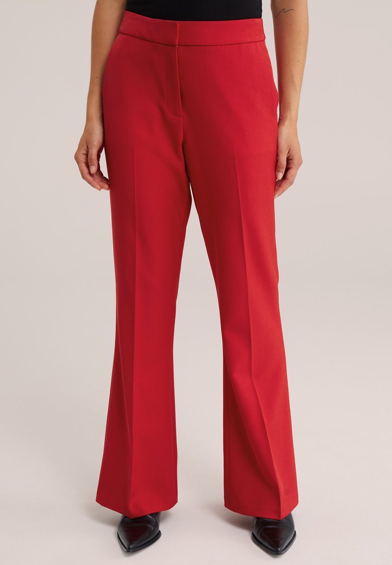Femme portant un pantalon rouge taille haute à jambes larges assorti à des chaussures noires à bouts pointus, debout devant un fond neutre.