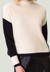 Pull en tricot côtelé noir et crème avec un col haut, présentant des manches longues en blocs de couleurs et un ourlet droit. Texture douce.