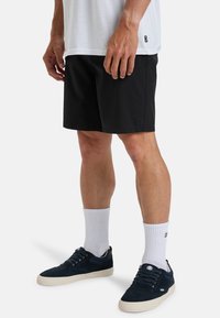 Černé kraťasy se hladkým povrchem, s malou nášivkou loga, v kombinaci s bílými ponožkami a tmavými teniskami. Zobrazena ležérní outfit.