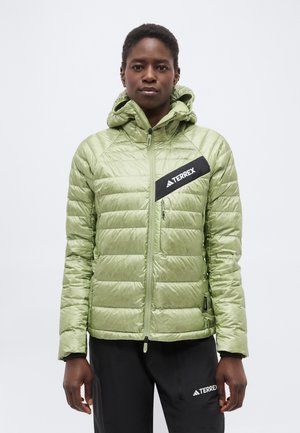 Henkilö, joka pukeutunut vaaleanvihreään tikattuun hupulliseen takkiin ja mustiin housuihin, molemmissa "TERREX"-logolla, seisoo yksiväristä taustaa vasten.