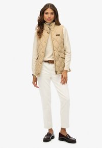 Gilet en beige matelassé avec fermeture à boutons-pression, poches latérales et col haut, associé à un haut crème et un pantalon blanc. Ceinture tressée marron et chaussures sombres.