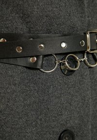 Grijze viltstof met een zwarte gestructureerde riem met metalen studs en ringen. Opvallende zwarte knoopdetail aan de linkerkant.