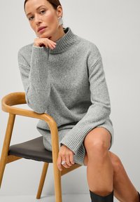 Pull en tricot gris avec col montant, poignets côtelés et coupe décontractée. Chaise en bois avec assise marron. Fond minimaliste.
