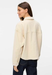 Beige fløjlsjakke med krave, lange ærmer og løs pasform. Har vandrette ribbede detaljer og knapper på ærmerne.
