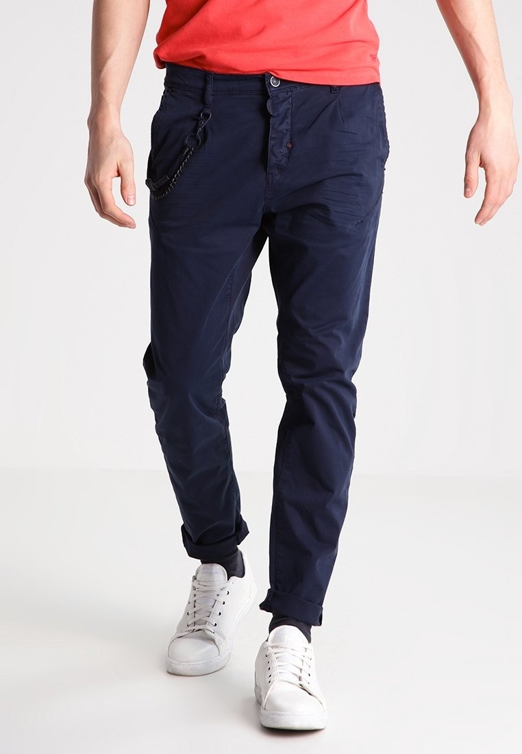 Homme portant un pantalon bleu marine retroussé, des baskets blanches et une chemise rouge, marchant sur un fond blanc uni.