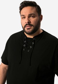 Muž s vousy, krátkými tmavými vlasy, oblečený v černé košili s šněrováním u výstřihu, dívající se mírně mimo kameru na bílém pozadí.