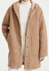 Abrigo de lana oversized en color marrón claro, con una superficie texturada y suave, capucha y dos grandes bolsillos frontales, abrochado con botones en la parte delantera.