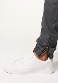 Białe skórzane sneakersy w połączeniu z szarymi spodniami z zamkami na nogawkach. Buty mają płaską podeszwę i okrągły czubek.