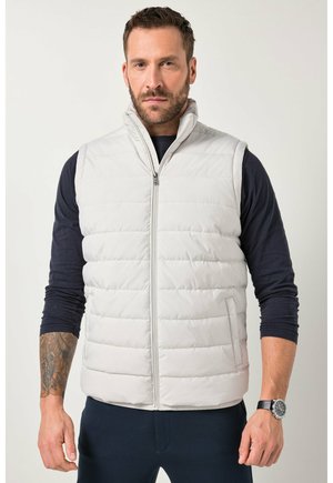 Mand med skæg iført hvid quiltet ærmeløs vest over sort langærmet trøje, stående mod neutral baggrund.