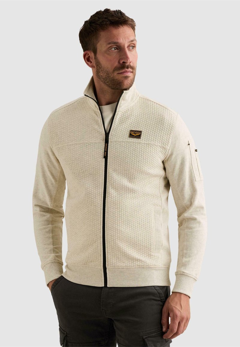 Homme portant une veste beige texturée à fermeture éclair avec un col haut et une fermeture noire, regardant vers sa gauche, mains dans les poches, sur un fond uni.