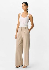 Beige broek met wijde pijpen, geplooide voorkant, hoge taille en een gladde textuur, gecombineerd met een aansluitend wit tanktop en minimalistische sandalen.
