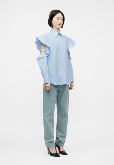 Femme debout portant un chemisier bleu clair à volants avec épaules dénudées, un jean large délavé clair et des escarpins noirs à bout pointu sur fond blanc.