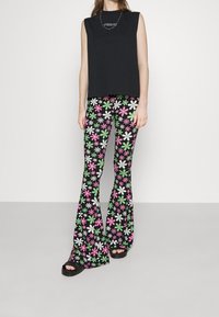 Vrouw in een zwart mouwloos topje, bloemenprint uitlopende broek in groen, roze, wit en zwarte sandalen tegen een witte achtergrond.