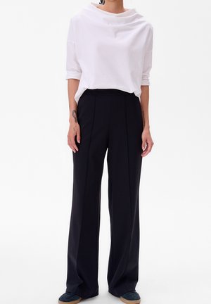 Personne portant un haut blanc ample avec manches retroussées et un pantalon noir taille haute à jambes larges, debout sur un fond uni.