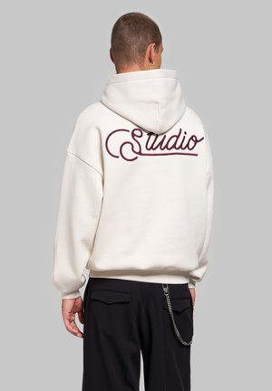 Personne portant un sweat à capuche blanc avec un texte "studio" bordeaux dans le dos et un pantalon noir avec un accessoire chaîne, vue de dos sur un fond gris.