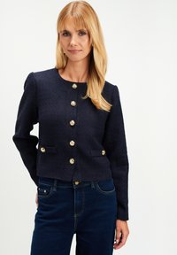 Kobieta z blond włosami ubrana w granatowy, strukturalny kardigan z guzikami w kolorze złotym oraz ciemnoniebieskie dżinsy na białym tle.