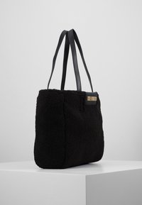 Svart fleece tote-väska med två svarta läderremmar. Har en rektangulär form och en guldig logotypaccent p å ena sidan. Mjuk textur.