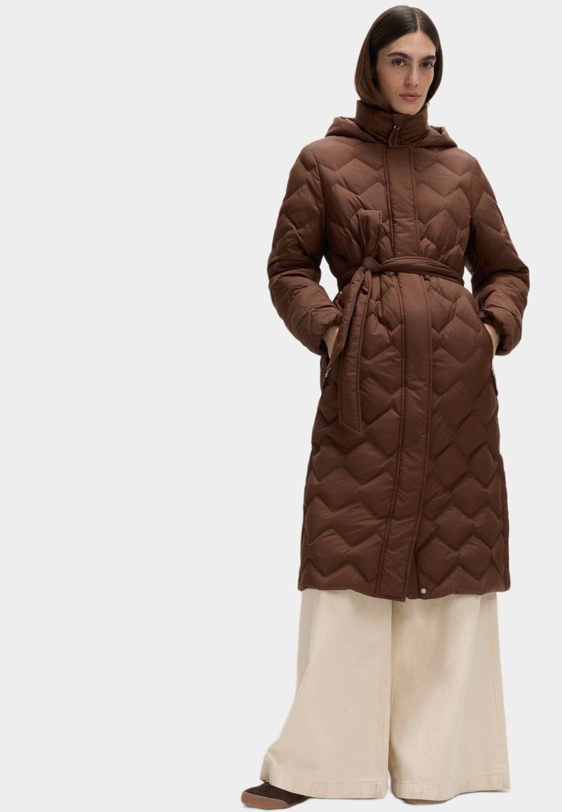 Brun quiltet lang jakke med hætte og bælte, med et diamantmønster og sidelommer, båret over cremefarvede vide bukser.