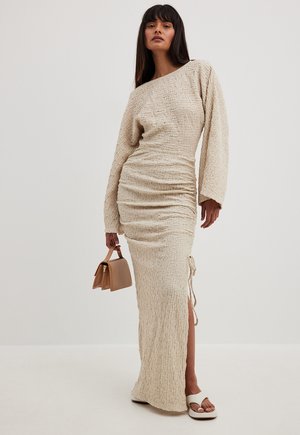 Robe beige, texturée, à manches longues avec des côtés froncés et une fente sur le côté. Associée à un petit sac à main beige et des sandales de couleur claire.