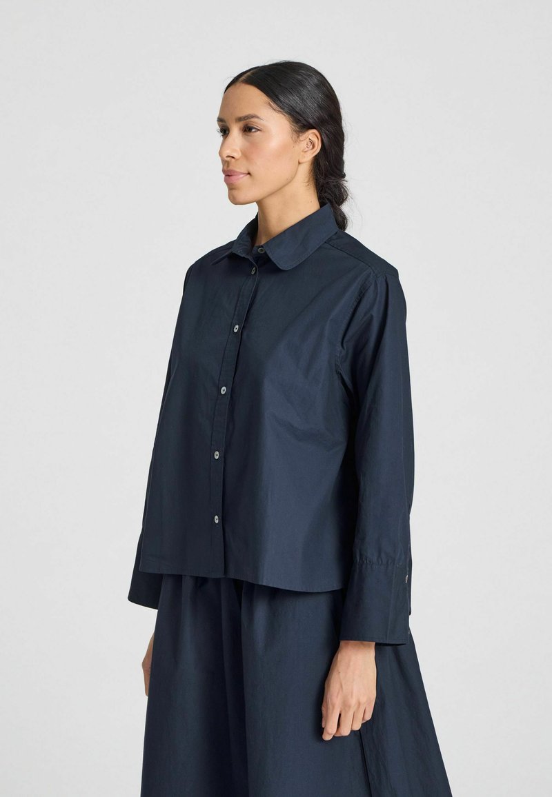 Camicia button-up blu navy con colletto strutturato, maniche lunghe e taglio ampio. Realizzata in tessuto liscio con una leggera lucentezza.