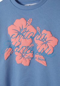 Világos kék pulóver, amelyet texturált rózsaszín hibiszkusz virágok díszítenek, és stilizált kék felirat olvasható rajta: "Aloha Honolulu."