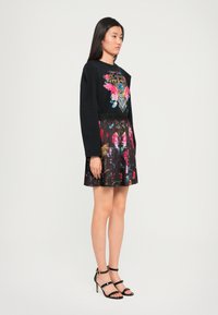 Top negro de manga larga con bordados florales coloridos, combinado con una falda plisada con patrones vibrantes sobre un fondo oscuro. Tacones negros con cintas.