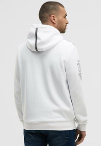 Sudadera blanca con capucha, fabricada con un tejido texturizado, que presenta rayas de acento negras en la capucha y un diseño impreso en la manga.