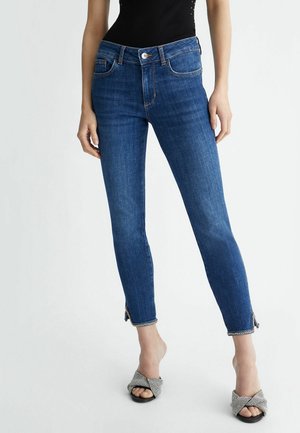 Nainen pukeutuneena sinisiin kapeisiin farkkuihin, joissa nilkanvetoketjut, ja mustavalkoruudullisiin avokkaisiin valkoisella taustalla.