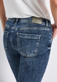 Tmavě modré džíny z denimu s vybledlou strukturou, obsahující dvě zadní kapsy a obdélníkovou štítek na pásku. Klasický design s pěti kapsami.