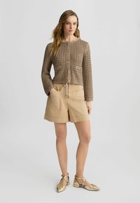 Klassisch geschnittene, strukturiert braune Strickjacke mit Knöpfen und zwei Vorder-Taschen. Kombiniert mit beigen Shorts und flachen metallischen Schuhen. Einfaches, modernes Design.