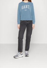 Felpa blu con la scritta "GANT 1949" in bianco, abbinata a jeans neri e sneakers grigie. La felpa ha maniche lunghe e un colletto arrotondato.