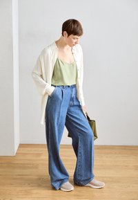 Top de satén verde claro, combinado con una camisa crema de botones, usado con jeans de pierna ancha azul de cintura alta y zapatillas beiges, sosteniendo un bolso verde.